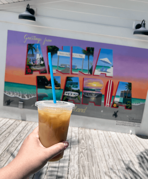 a hand holding a cup of iced coffee with a postcard inspired mural in the background